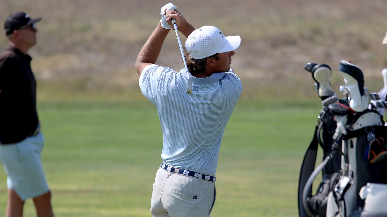Golfer mid-swing on a golf course, wearing a light blue polo and white cap, with a bag of clubs nearby.