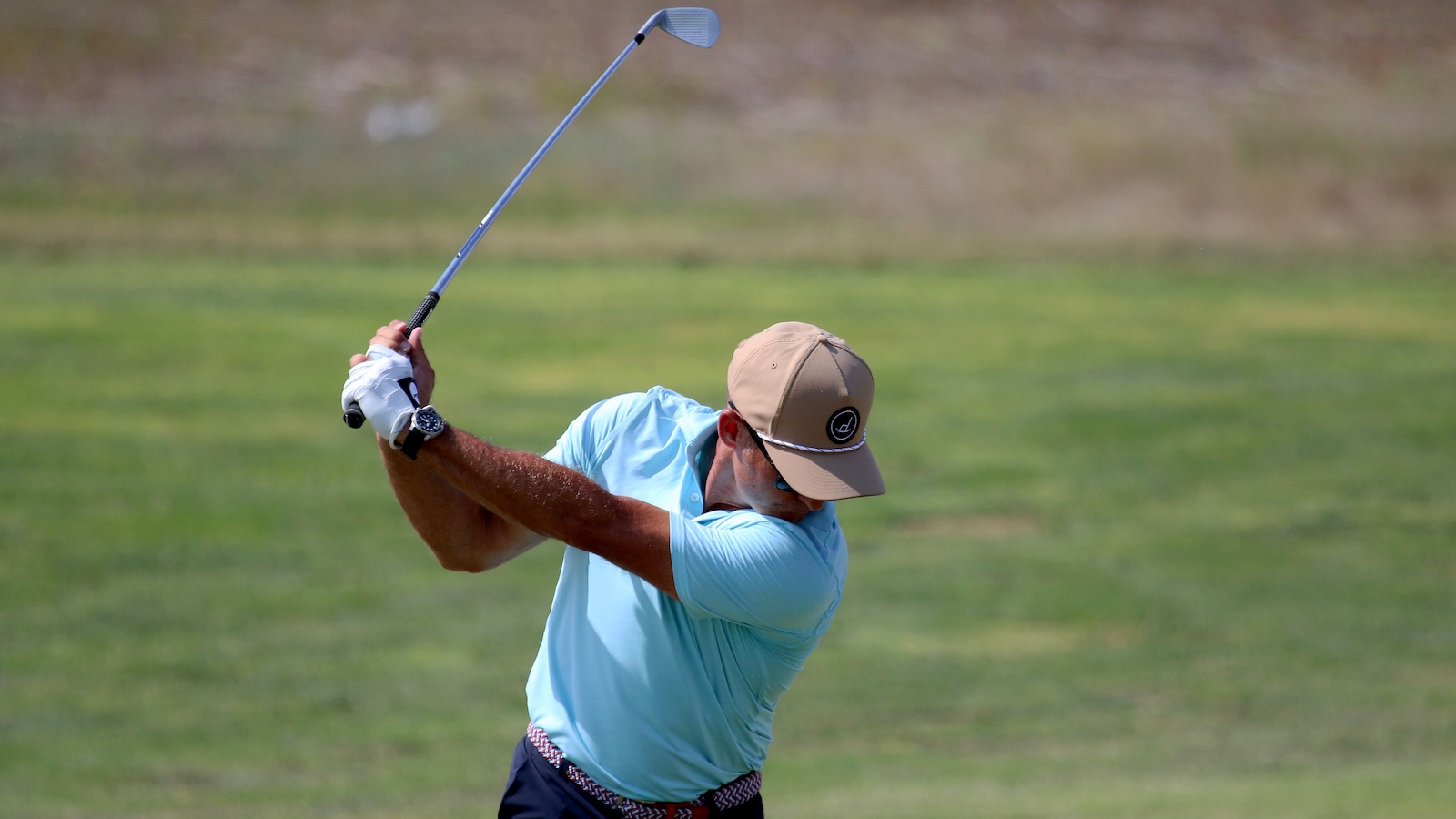 Golfer in a light blue shirt swings a golf club on a grassy course, wearing a tan cap and white glove.