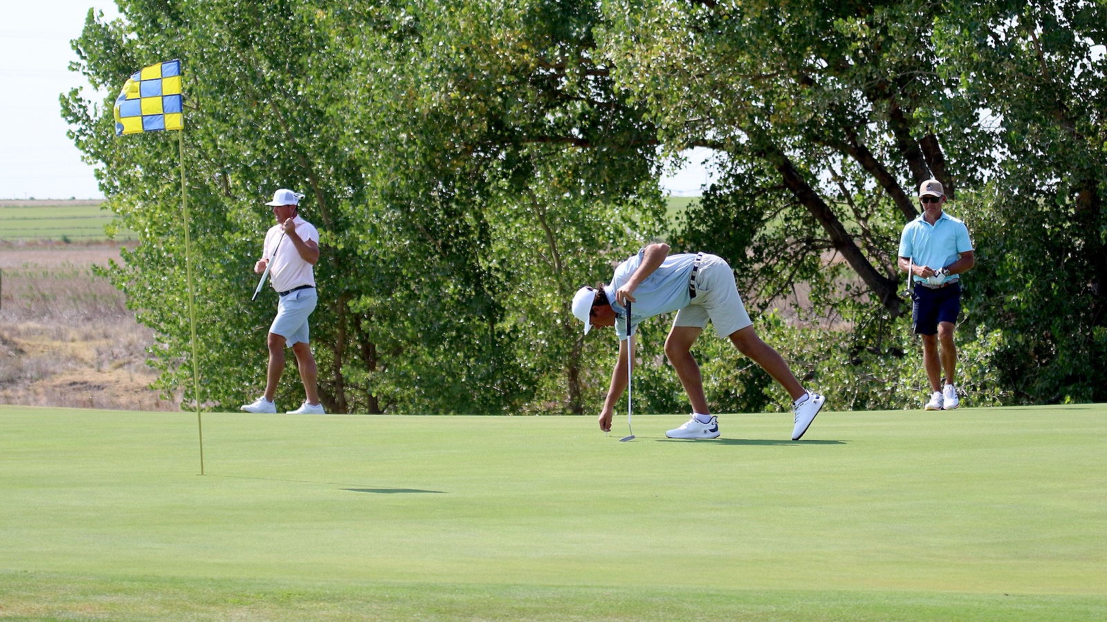 Golfer lining up a putt on a green with a two-tone checkered flag nearby while two players watch under trees