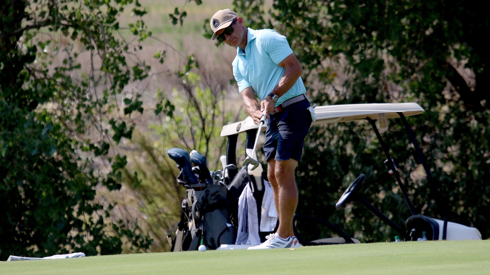 Man in a light blue polo and cap tees off on a golf course, gripping a club near a golf bag and trolley in the background.