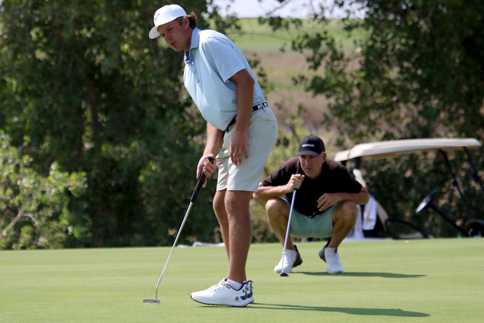 Golfer in a light blue shirt and white cap lines up a putt on a green while a teammate crouches behind with a club; a golf cart is in the background.