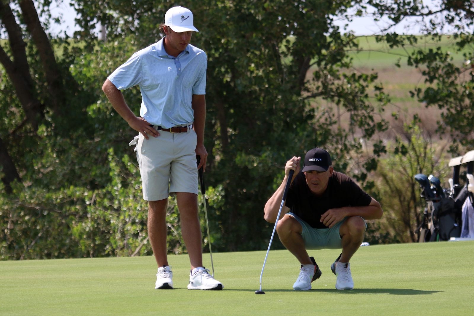 Two golfers on a green: one standing with hands on hips while the other crouches with a putter ready to line up a shot.