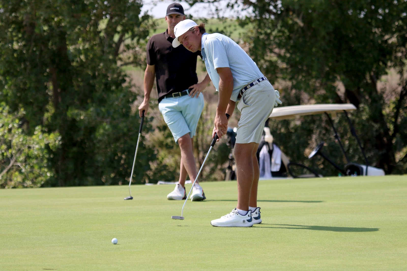Two men playing golf on a green putting surface, one bending to line up a putt as the other watches nearby.