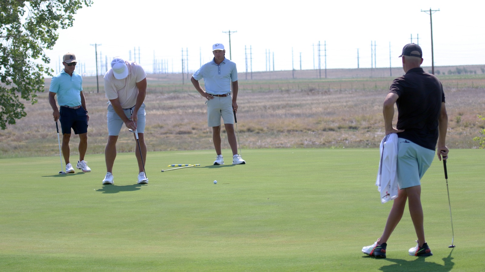Four golfers on a sunny course, one bending to putt while three others watch nearby.