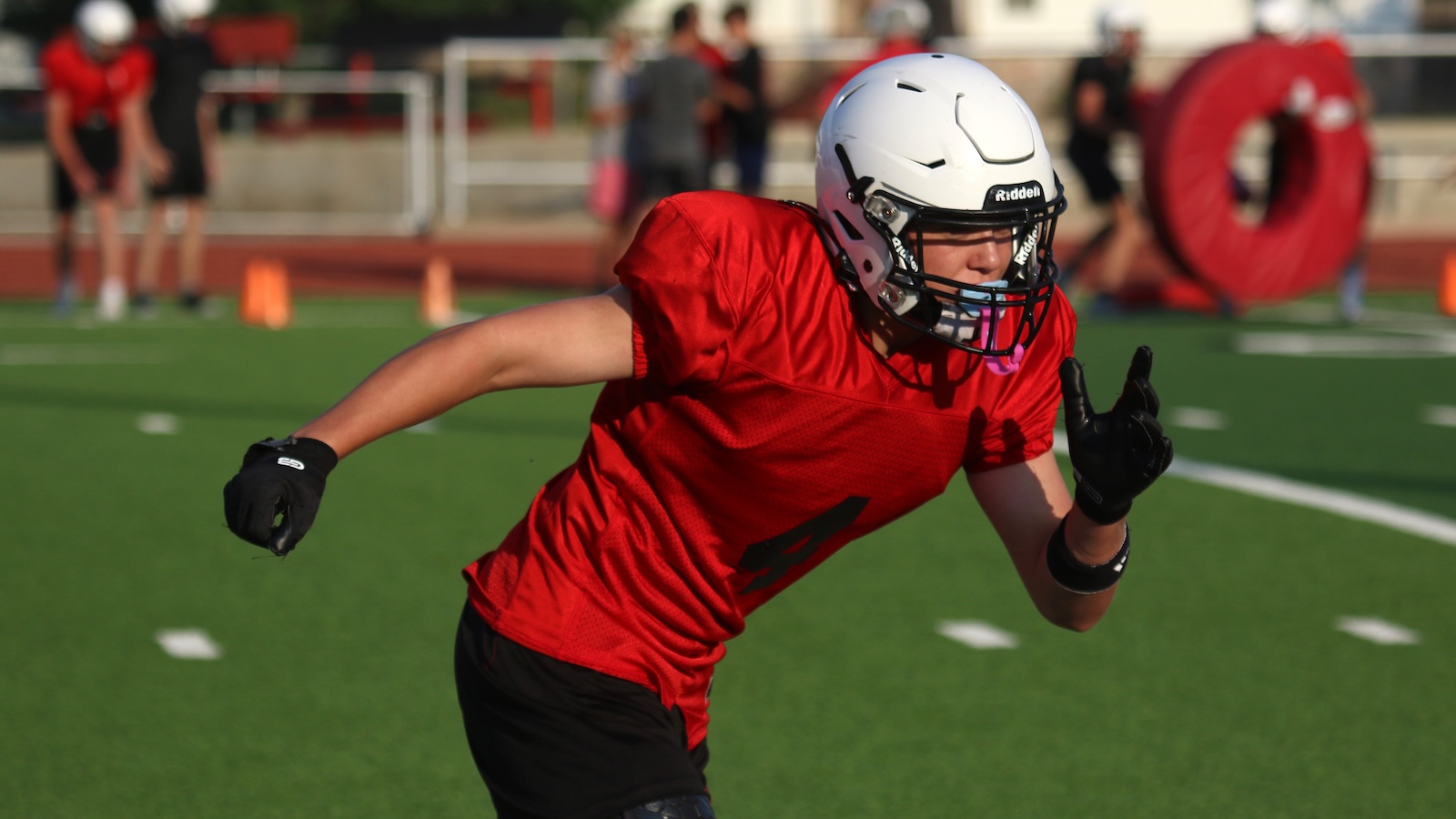 Football player in a red jersey and white helmet sprinting on a green practice field with cones in the background.