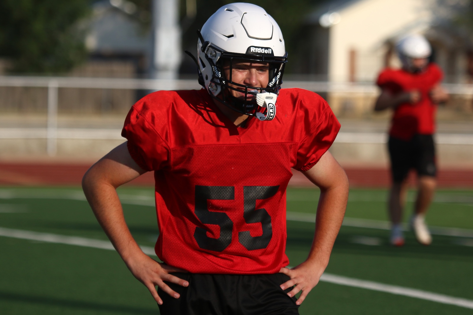 Football player in a red jersey with number 55 stands with hands on hips on a green turf field, wearing a white helmet and mouthguard.