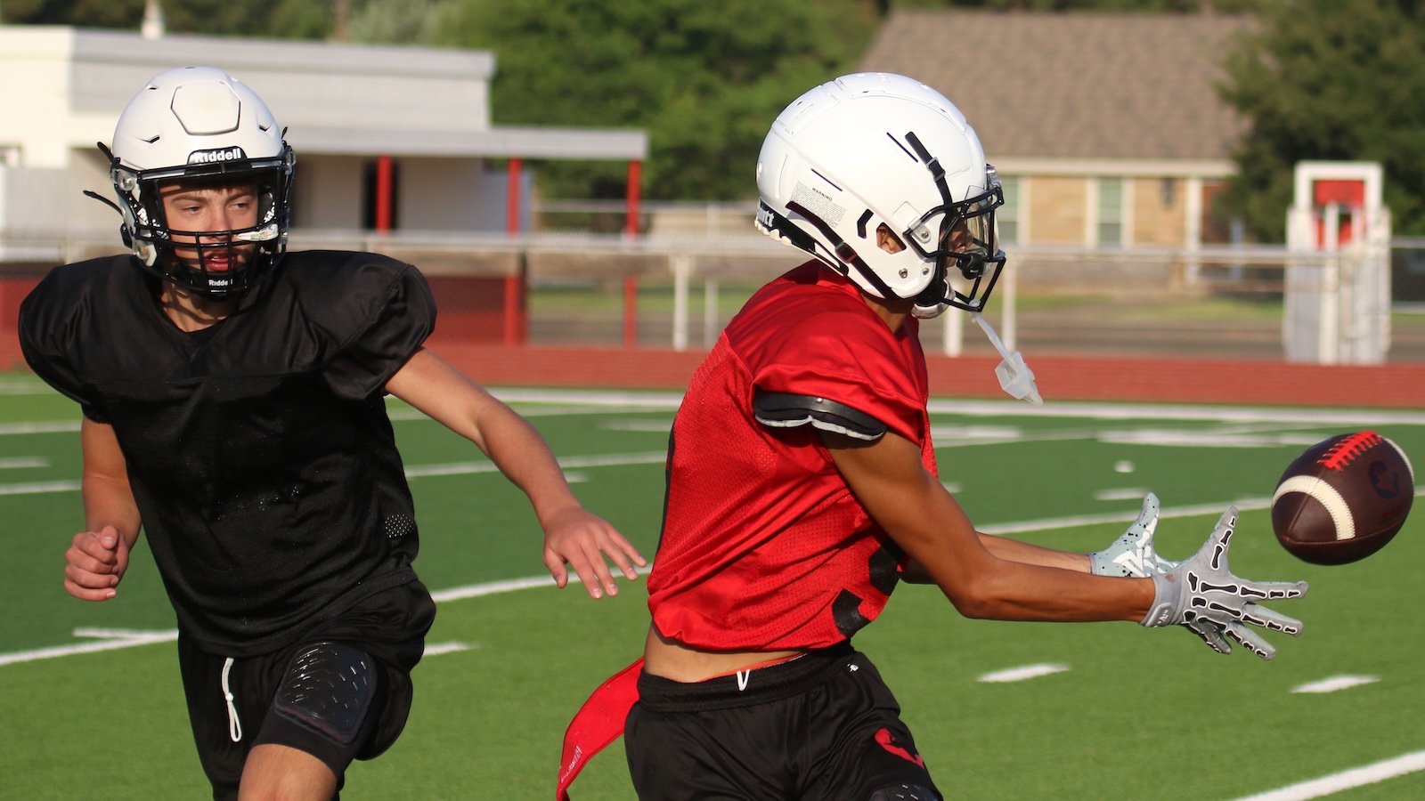 Two football players wearing helmets race on a green field, red jersey player reaching for a loose football as the black-jersey teammate pursues him.