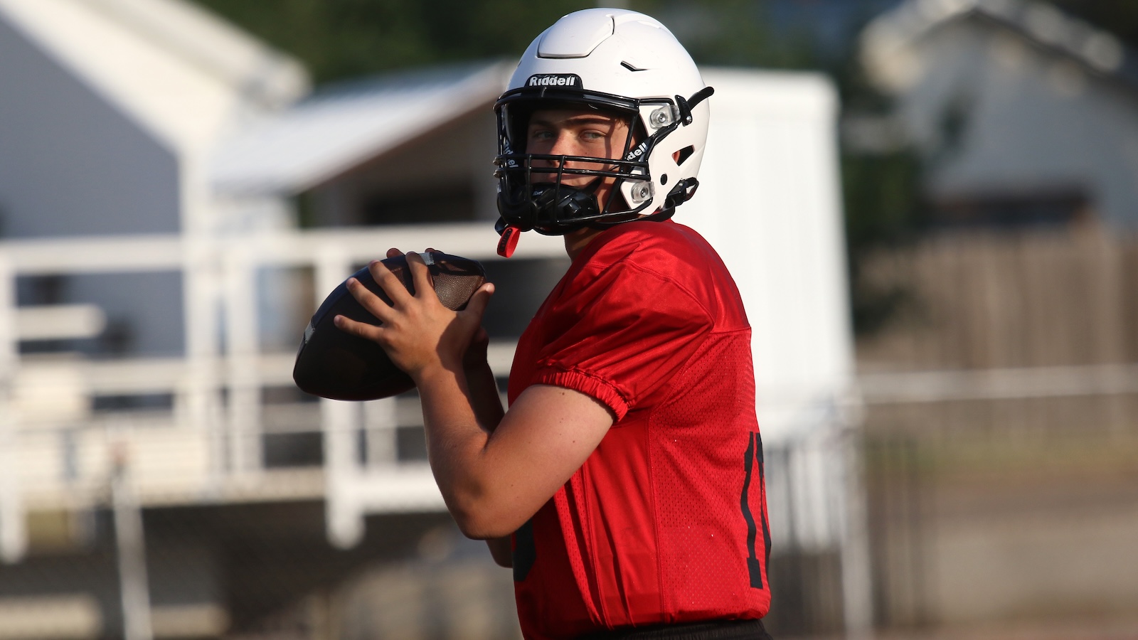 Quarterback in a red jersey and white helmet holds a football, ready to throw on an outdoor field.