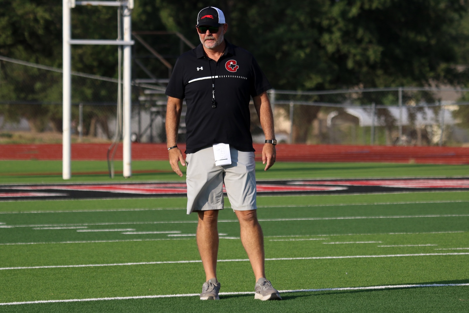 Coach standing on a football field in a black polo, gray shorts, cap, sunglasses, and a whistle on a lanyard around his neck.”