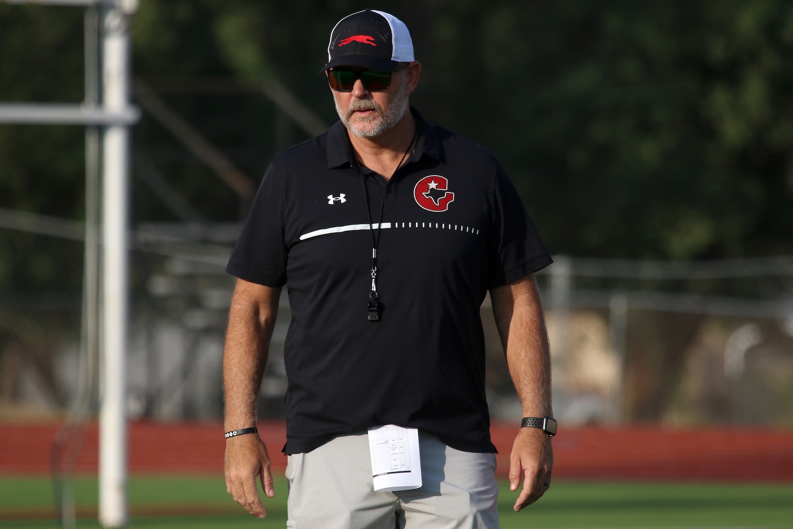 Athletic coach standing on a field wearing a black polo with a red emblem, sunglasses, and a cap, with a whistle around his neck and a notebook clipped to his shorts.