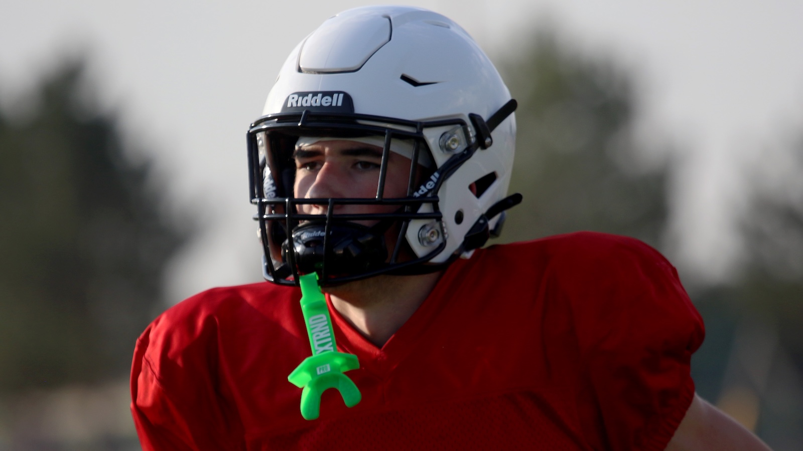 American football player wearing a white Riddell helmet and red jersey, looking focused on the field with a bright green mouthguard hanging from his neck.