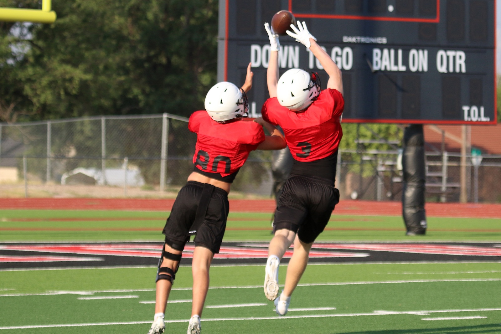 Two football players in red jerseys jump and reach for a ball near the end zone as a scoreboard looms in the background.