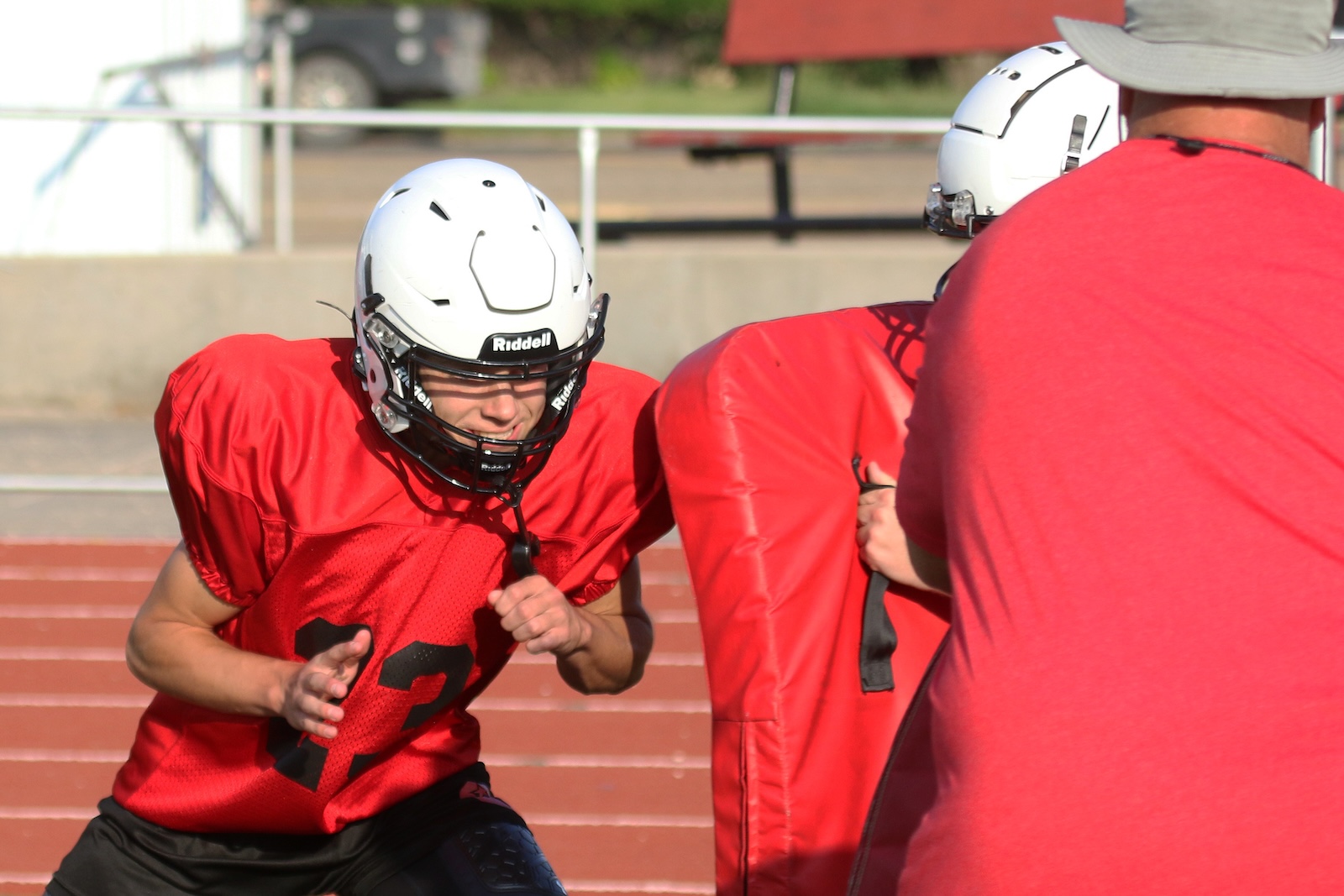 Football players in red jerseys practice tackling drills on a field, with a padded dummy and a coach nearby