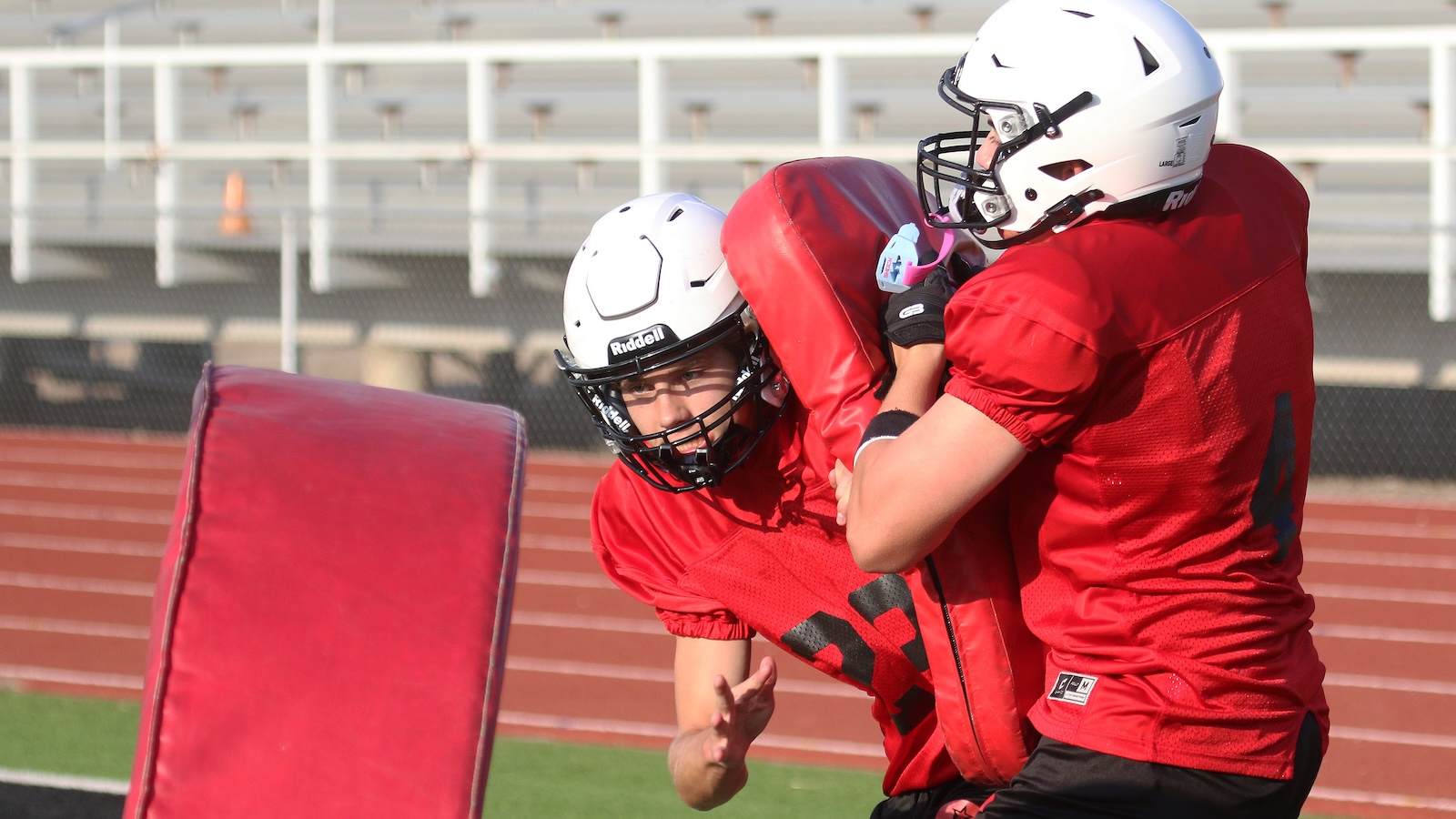 Two football players in red practice jerseys and white helmets drill blocking drills with a padded shield on an outdoor field.