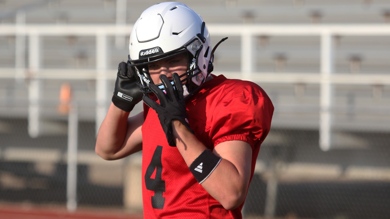 American football player in a red jersey number 4 adjusts his helmet on a practice field.