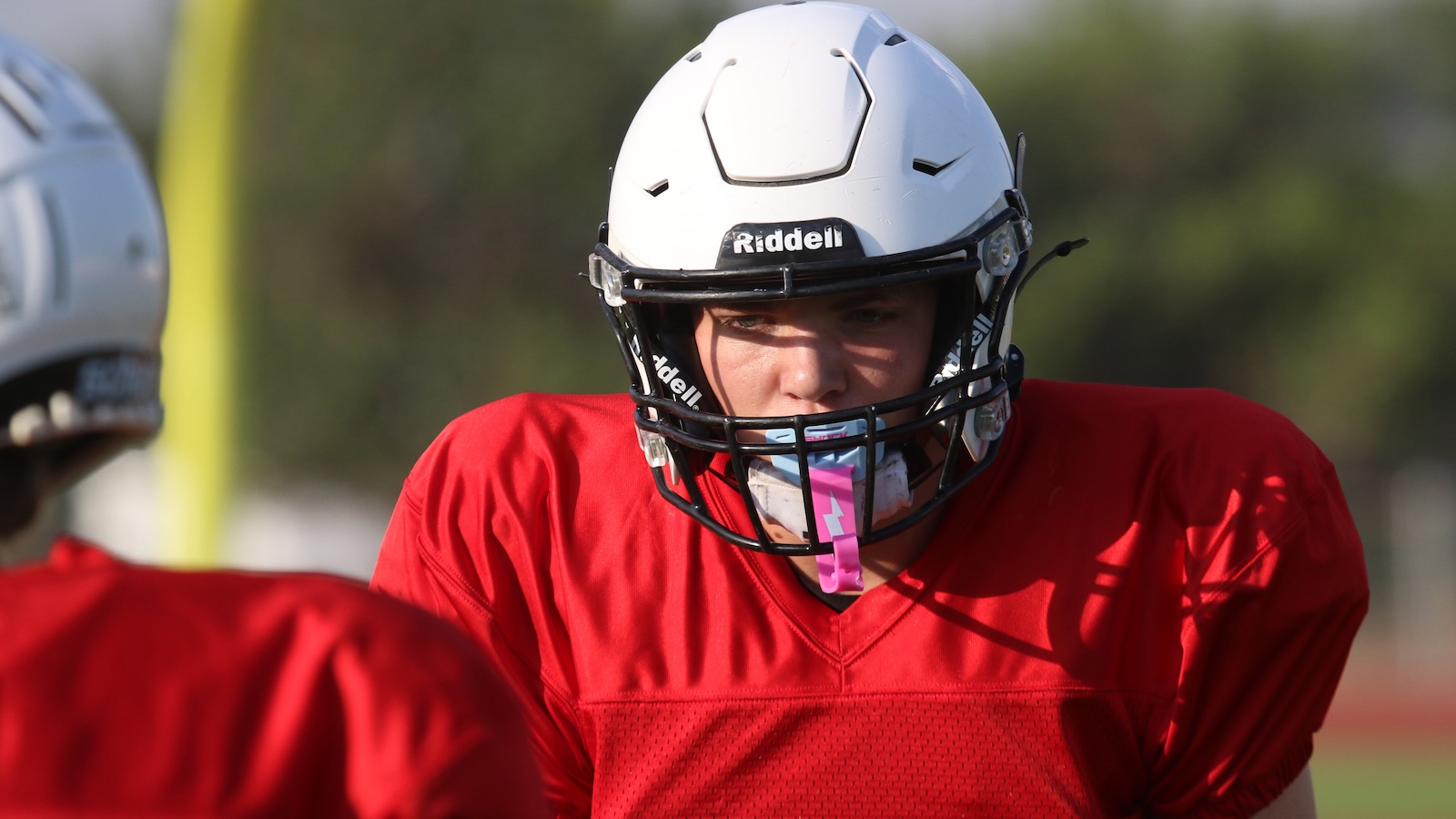 Football player in a red jersey and white Ridell helmet facing another player on the practice field.