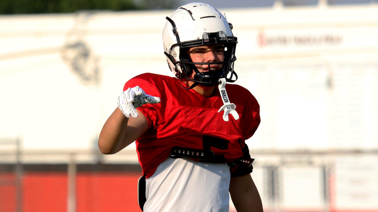 Football player in red jersey pointing with gloved hand during practice on a sunny field, wearing a white helmet and protective gear
