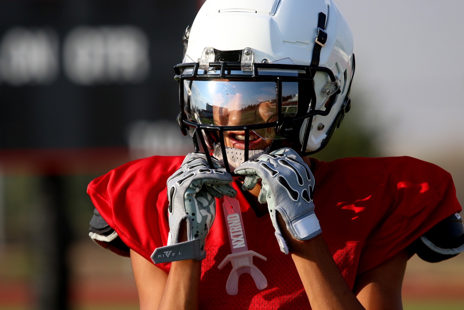 Football player in a red jersey adjusts a mouthguard while wearing a white helmet with a face mask on the field.