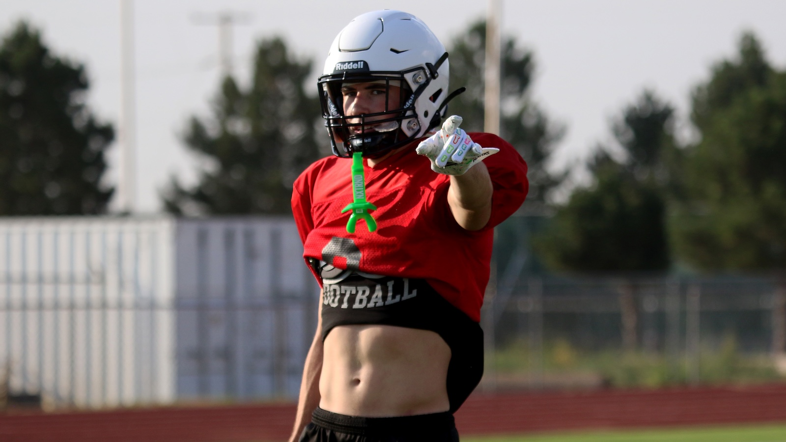 Football player in a red practice jersey and white helmet points forward on a field during football practice with a green mouthguard hanging from the jersey area.