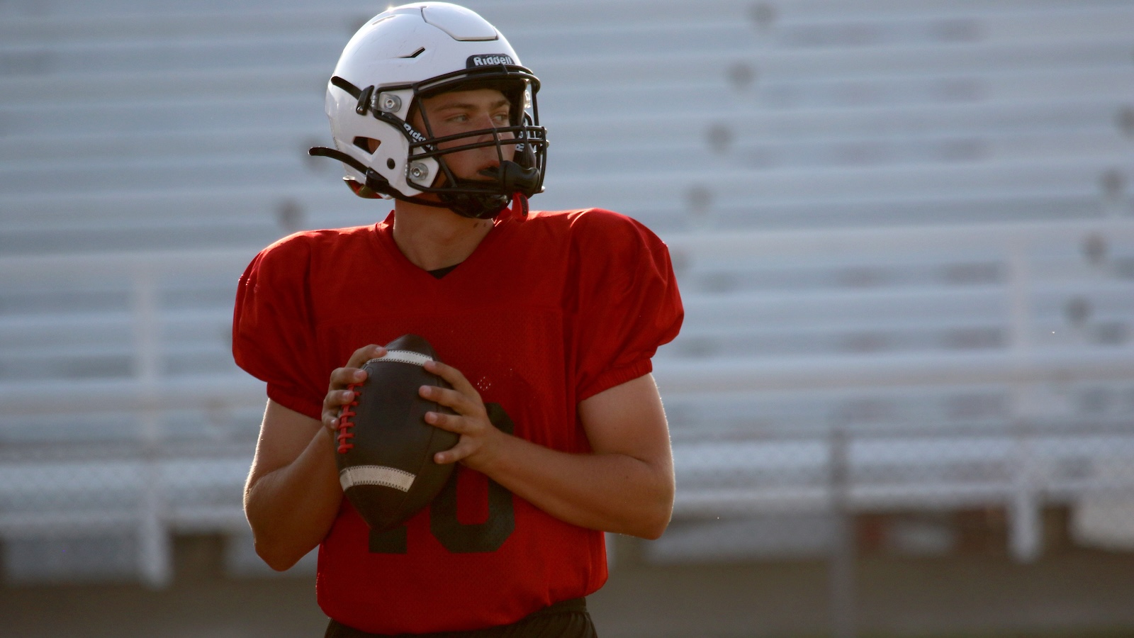 Quarterback in a red jersey (number 10) holding a football, ready to throw, wearing a white helmet on a football field in stands background.