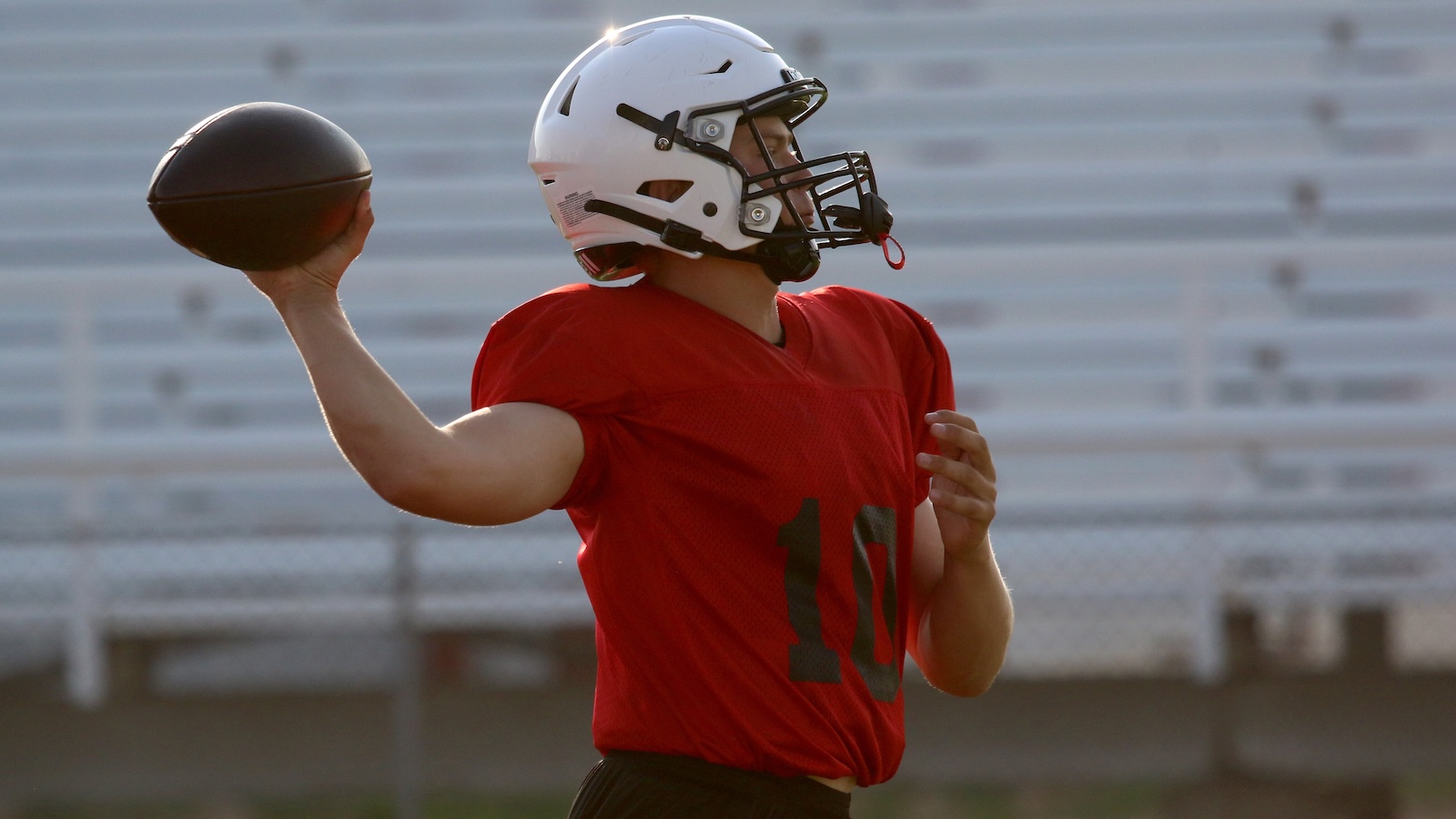 Quarterback in a red jersey with number 10 throwing a football on a practice field wearing a white helmet.