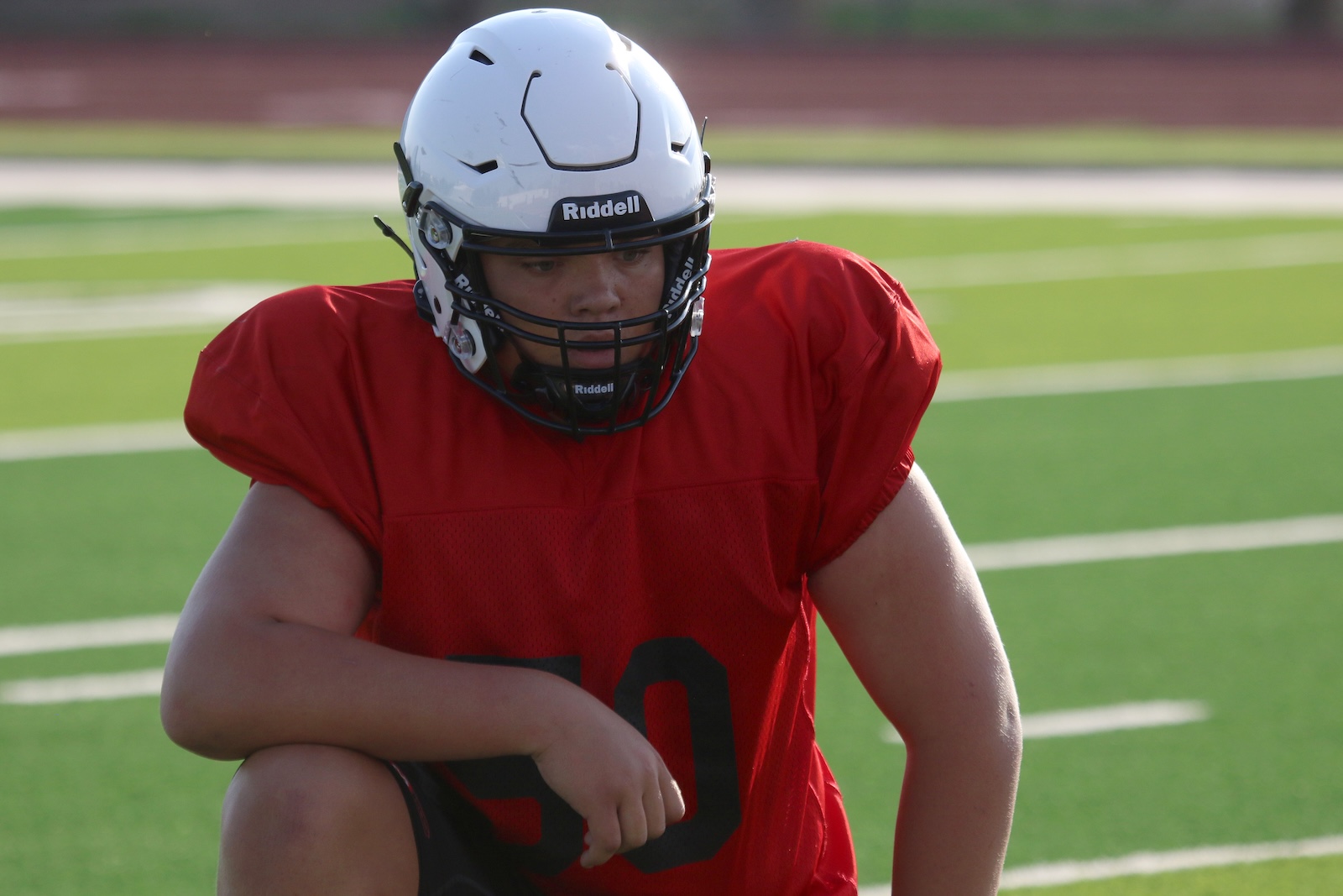 Football player in a red jersey and white helmet on a bright green field, squatting with one knee up and looking ahead.