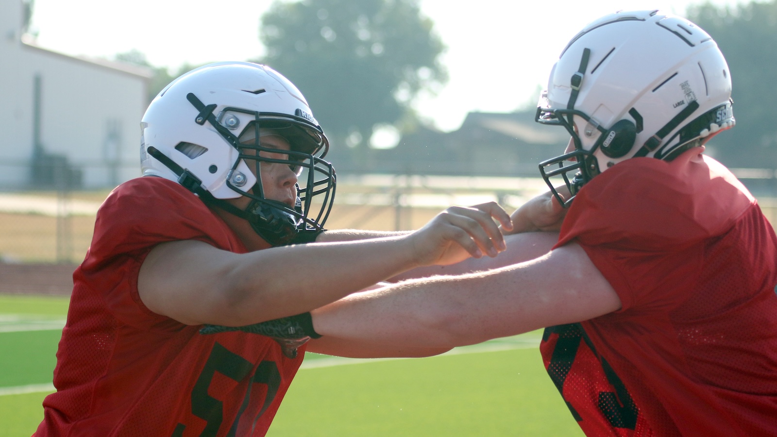 Two football players in red jerseys practice a blocking drill, gripping each other’s shoulders while wearing white helmets on a sunny field.