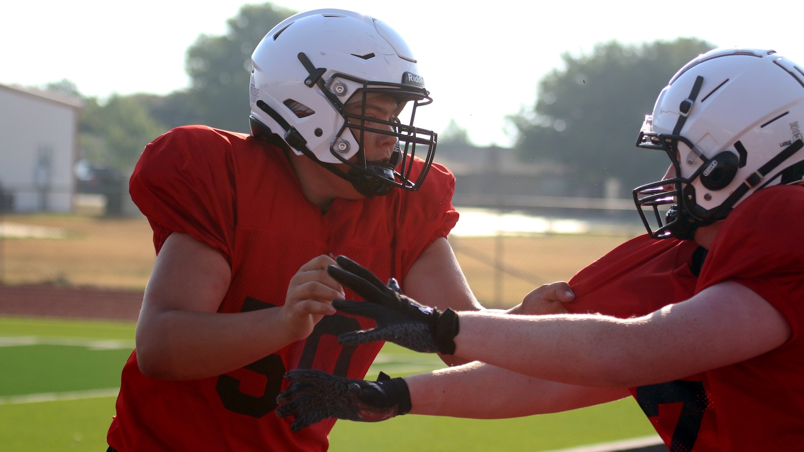 Two football players in red jerseys and white helmets engage in a drill on a sunny practice field, hands ready for contact