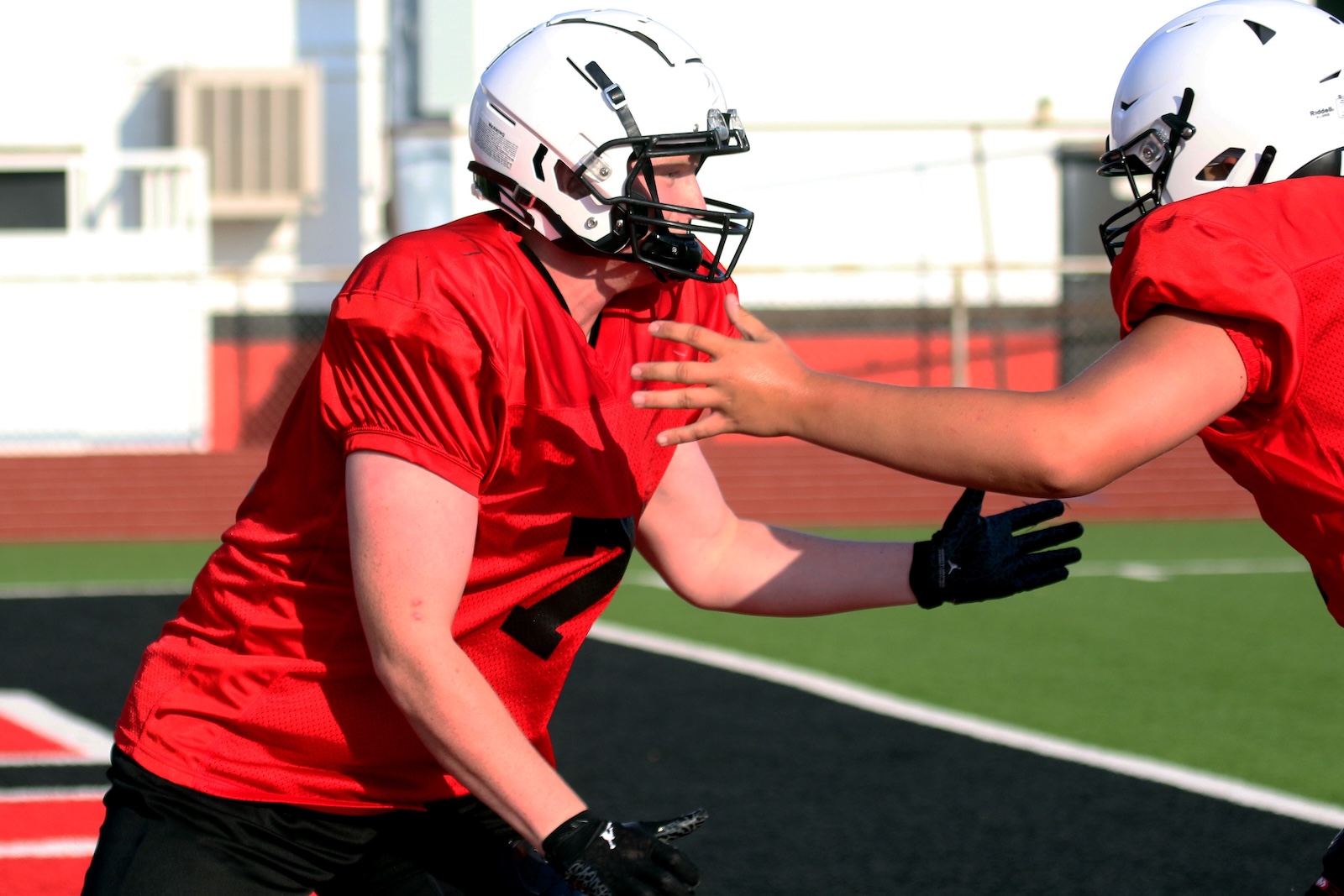 Two football players in red jerseys and white helmets practice blocking on a football field with artificial turf.