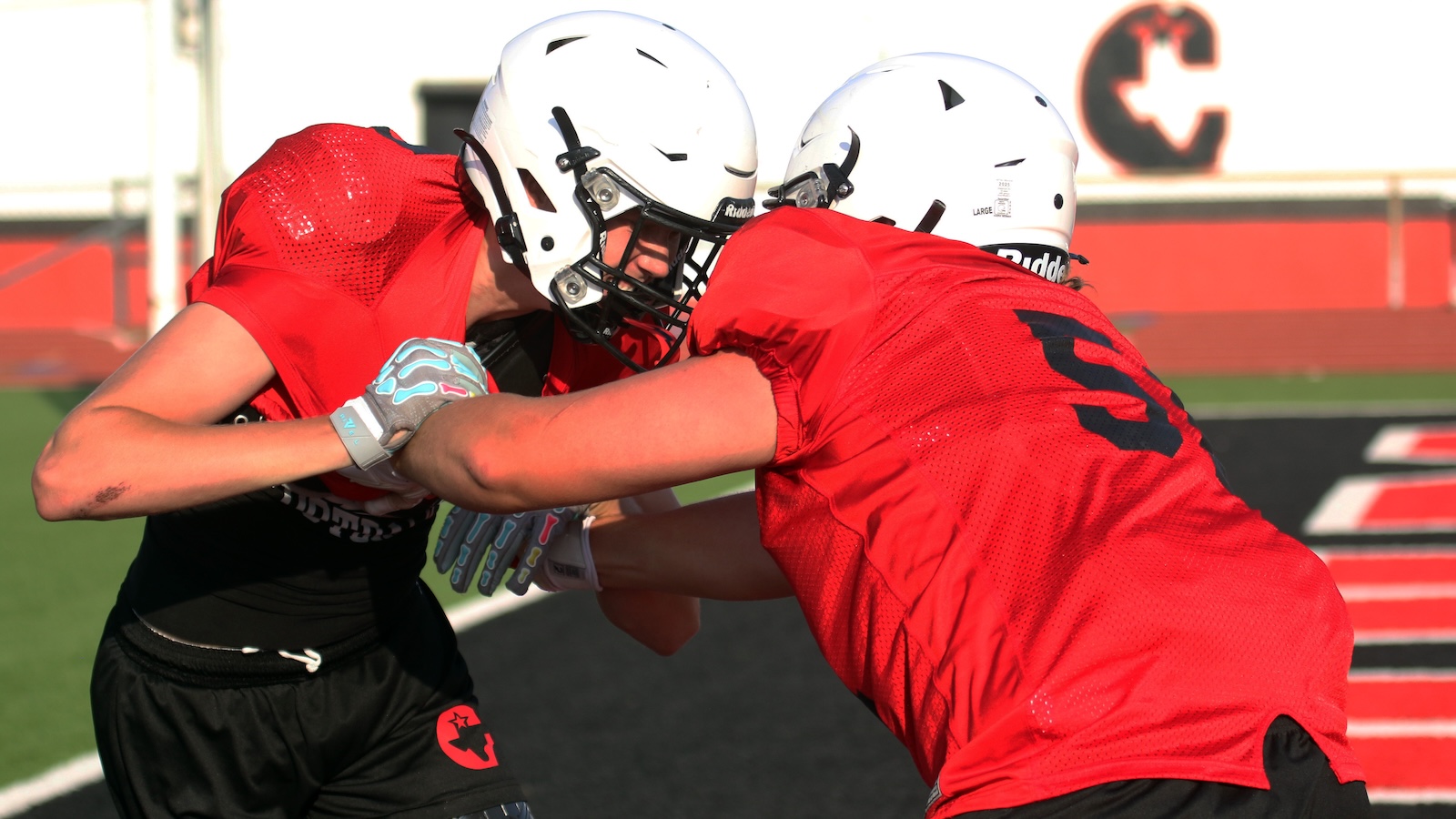 Two football players in red jerseys and white helmets lock arms in a tackling drill on a green field with a track in the background.