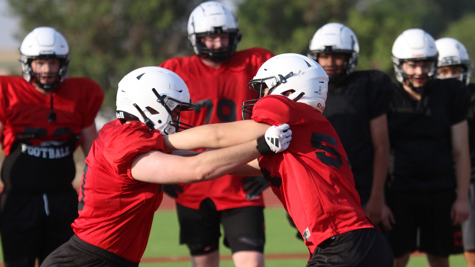 Two football players in red jerseys engage in a blocking drill on a green field while teammates watch in the background.