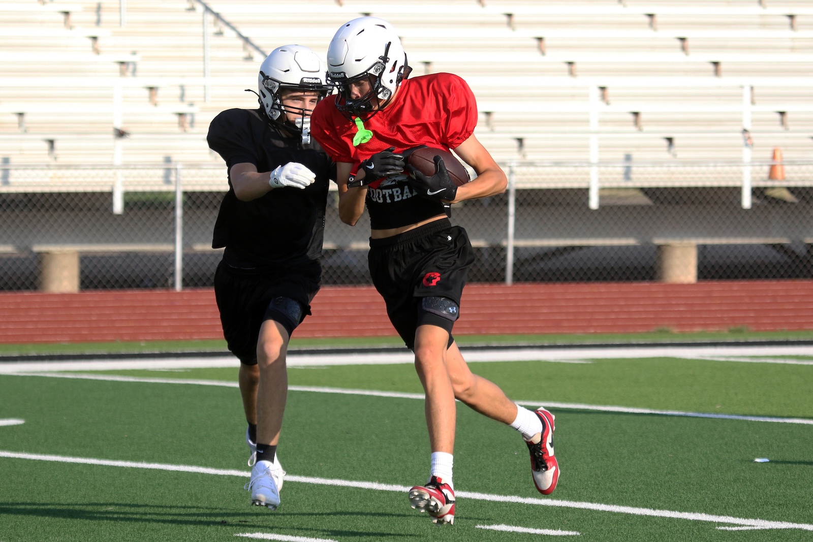 Two high-school/college football players sprint on a turf field, red-jersey runner carrying the ball while a black-jersey defender pursues behind him.