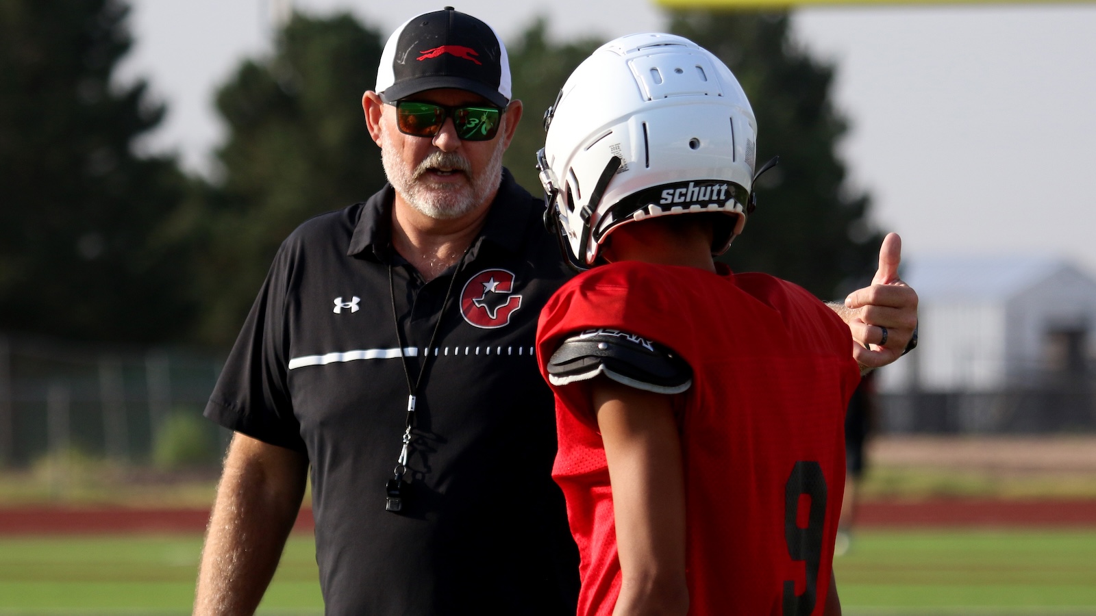 Football coach in sunglasses speaks to a player in a red jersey on the practice field, whistle at neck.