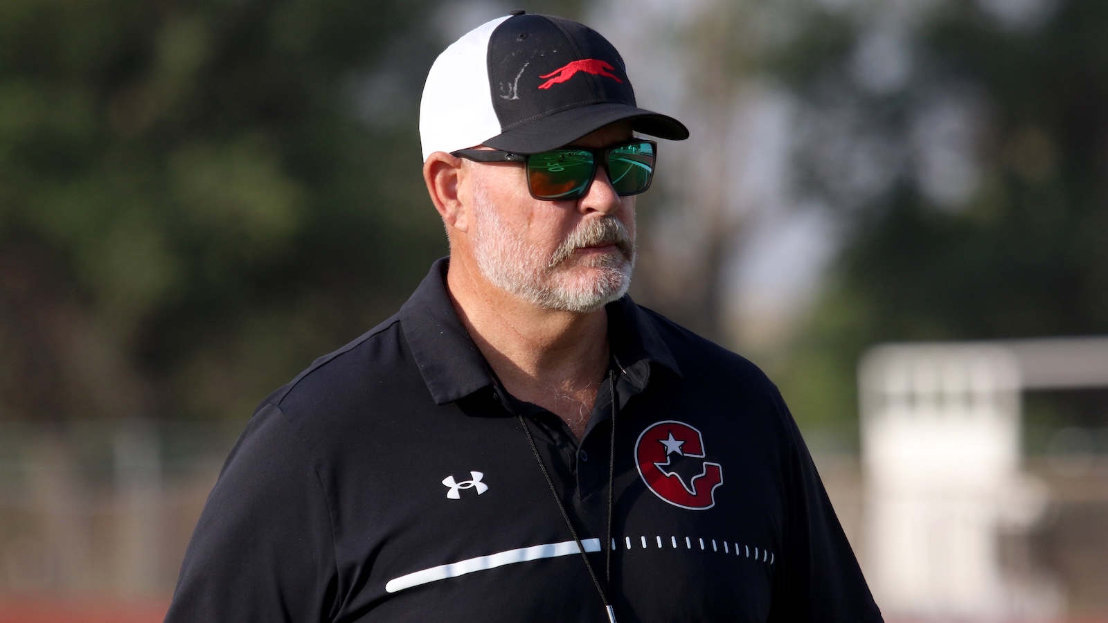 Older man with gray beard wearing a black cap, sunglasses, and a black polo with a red logo, standing outdoors.