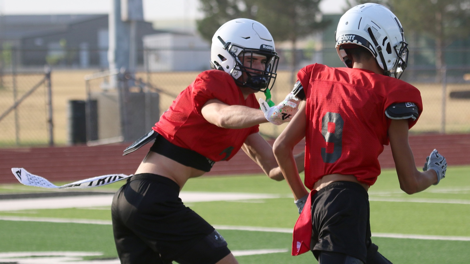 Two football players in red jerseys and white helmets practice on a turf field, colliding in a drill with a fence in the background.