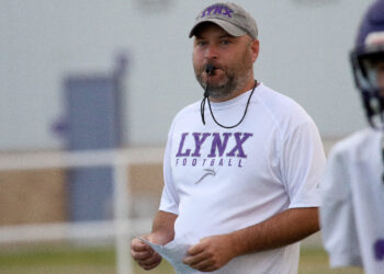 Spearman - HC Brandon Reagan 2 | Press Pass Sports Football coach wearing a white LYNX Football shirt with a whistle around his neck, holding play sheets on a practice field