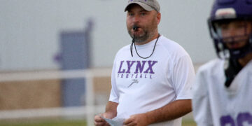Football coach wearing a white LYNX Football shirt with a whistle around his neck, holding play sheets on a practice field
