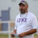 Spearman - HC Brandon Reagan 2 | Press Pass Sports Football coach wearing a white LYNX Football shirt with a whistle around his neck, holding play sheets on a practice field