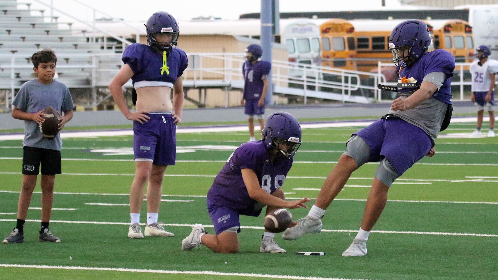 Youth football practice: a player kneels to place a ball as another player in purple jersey attempts a tackle on the ball carrier on a green synthetic field.