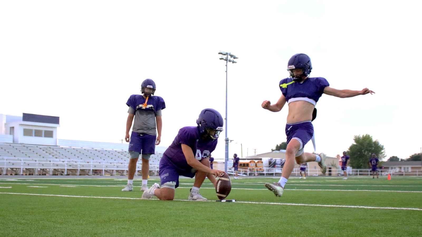 Young football players in purple uniforms on a field; one kneels to snap the ball while a teammate runs forward in a purple jersey.