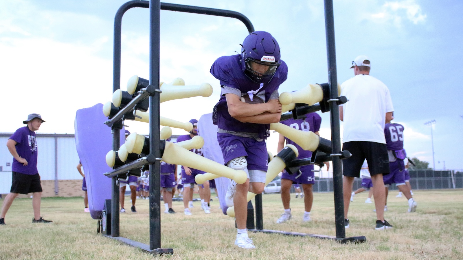 Youth football player in purple uniform and helmet crawls through a hurdle-like obstacle with yellow foam poles during practice on a grass field.