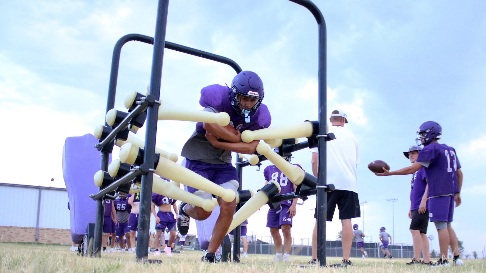 Youth football players in purple uniforms practice agility, crawling through a foam-padded obstacle on a grassy field during drills.