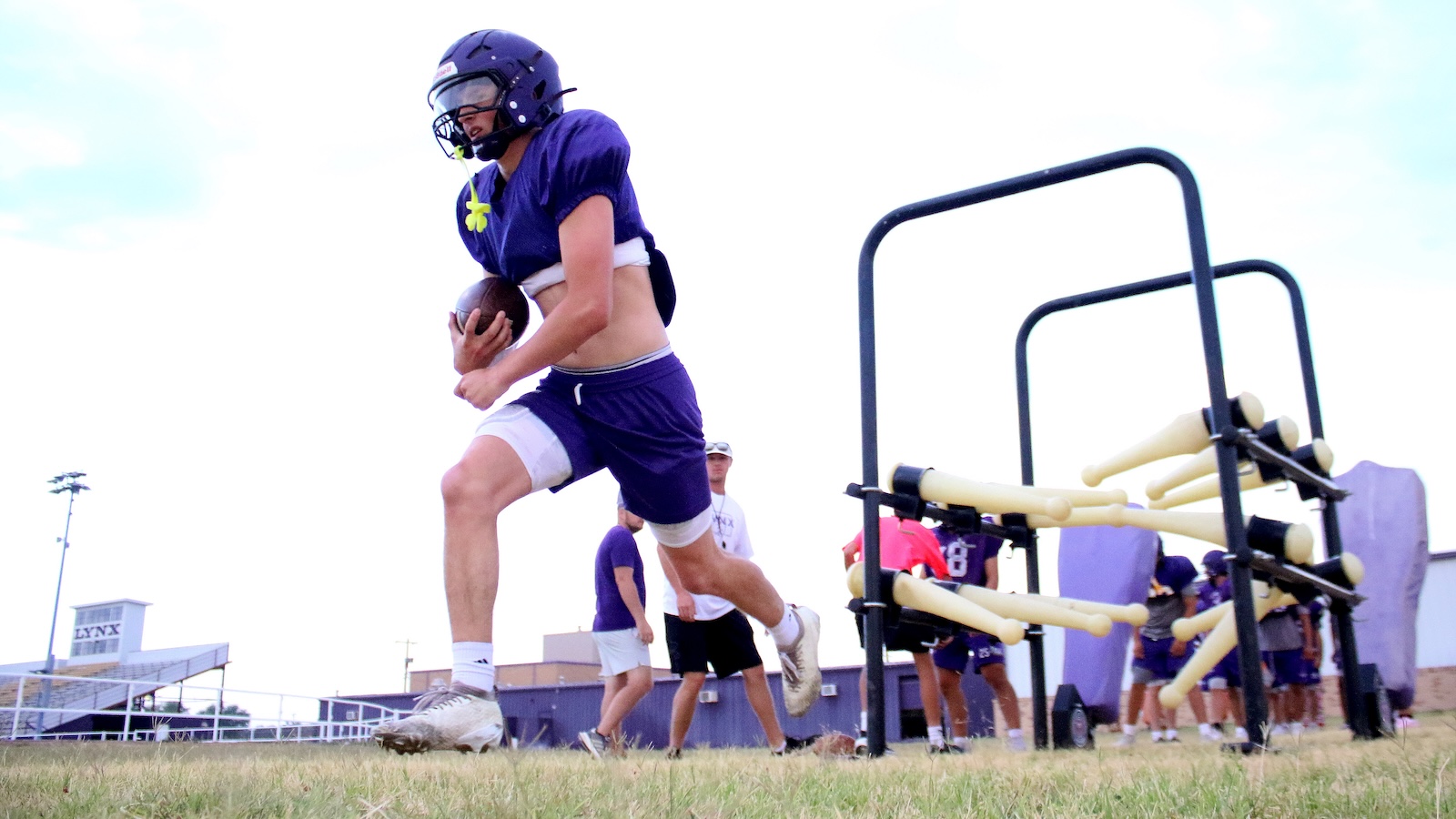 Football player in purple uniform runs with ball during practice on a grassy field, with training dummies to the right.