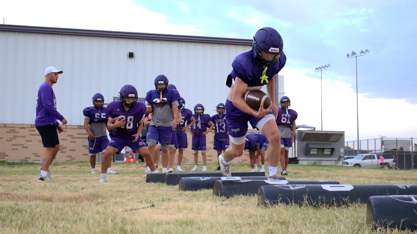 Young football players in purple uniforms practice sprinting through obstacle pads on a sunny field as a coach watches nearby.