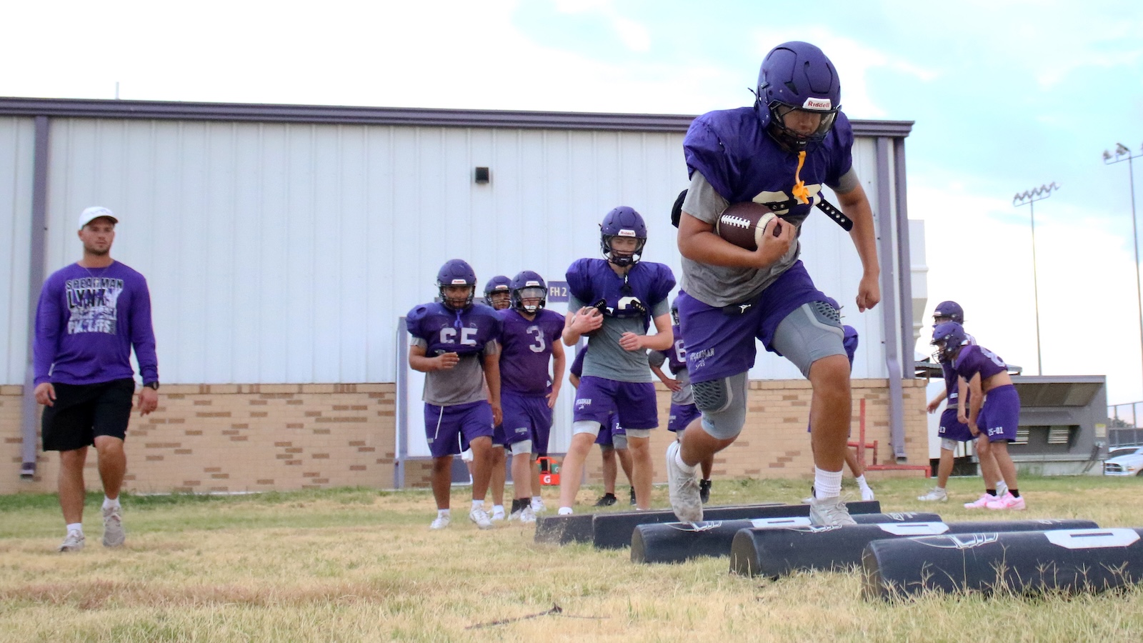Football practice: a player in a purple uniform sprints over agility pads while holding a ball, teammates and coach in the background
