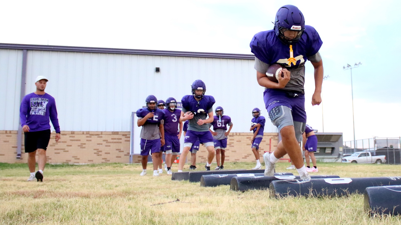 Youth football practice: players in purple uniforms run drills on artificial pads on a grassy field, coach nearby.