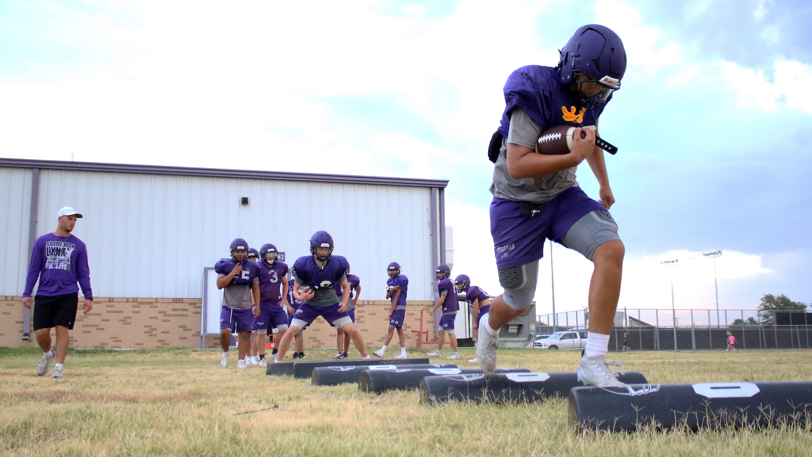 Young football players in purple practice jerseys sprint over padded blocking sleds on a grassy field, coach watching nearby.