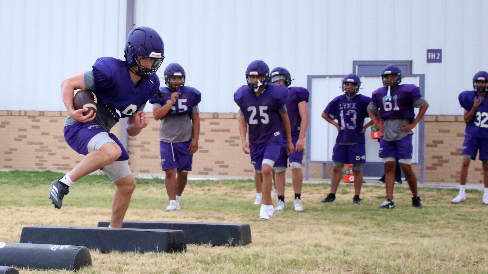 Young football player in purple jersey runs with ball over padded obstacles during practice.