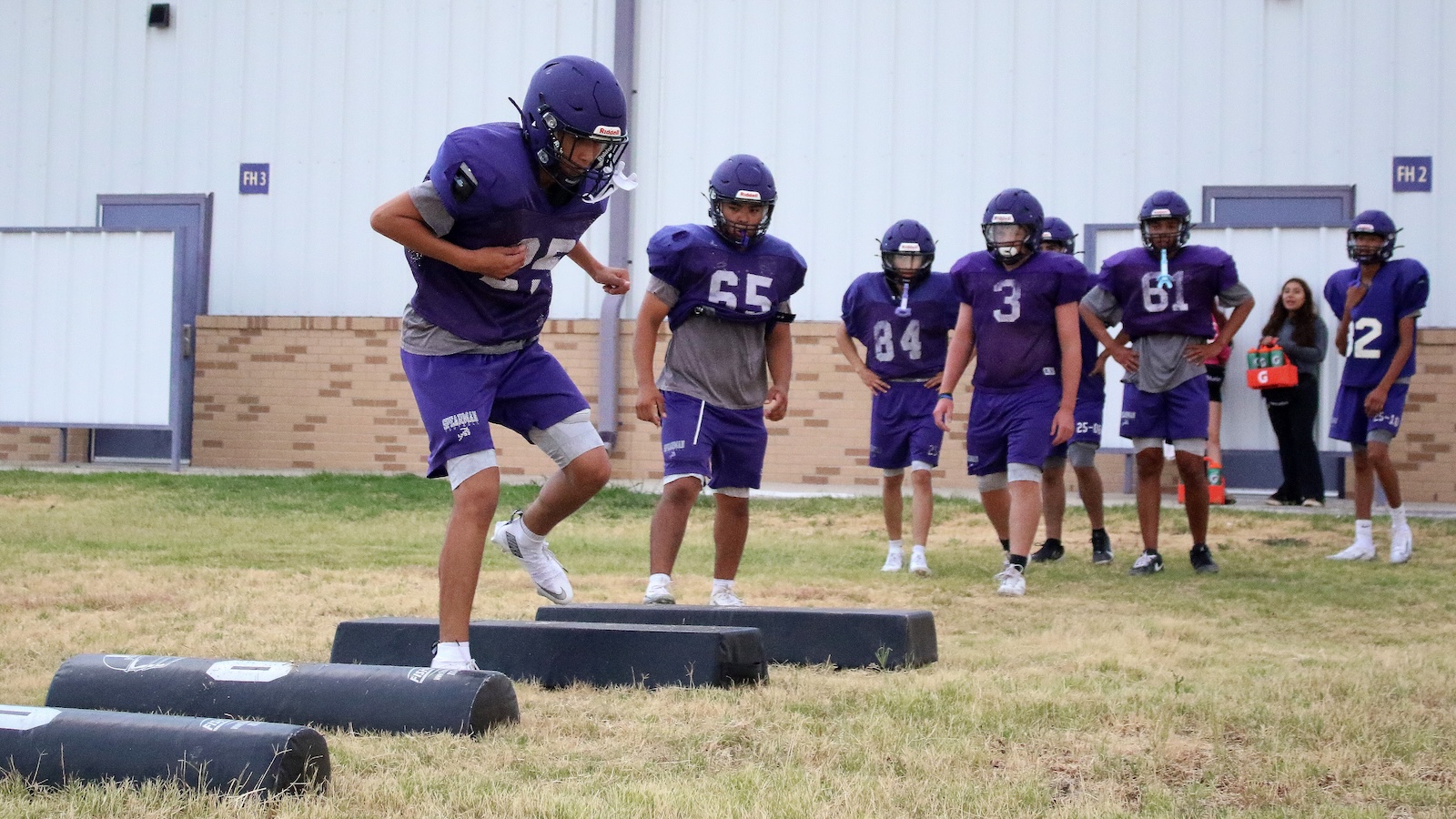 Youth football players in purple uniforms drill agility on a grassy field, stepping over black foam pads as teammates watch.