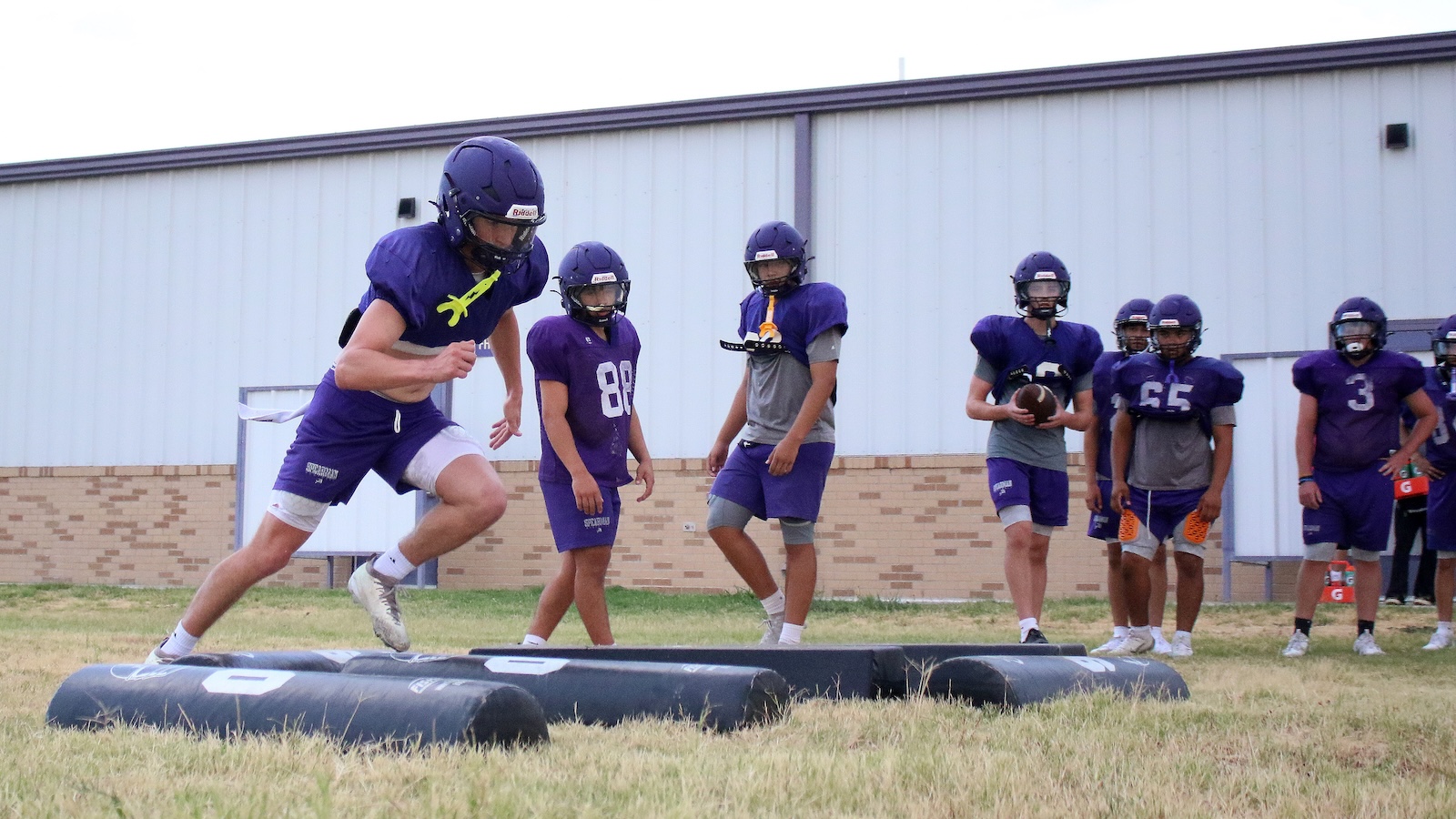 Youth football players in purple uniforms watch as a teammate sprint-drills over a padded hurdle on a grassy field.